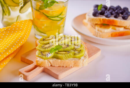 Rahm Käse und Obst Sandwiches. Sandwiches mit Frischkäse, Kiwi, Blackberry und Banane auf ein Holzbrett und Gurke Limonade in einem Glas Stockfoto