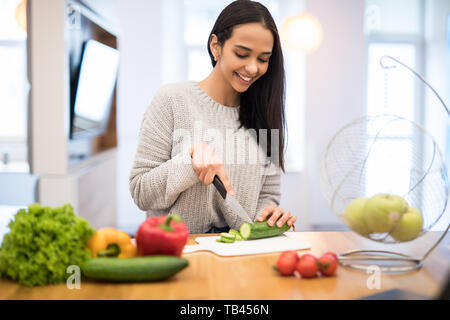 Die junge Frau schneidet Gemüse in der Küche mit einem Messer und Laptop auf dem Tisch. Salat. Diät. Diät Konzept. Gesunde Lebensweise. Cookin Stockfoto