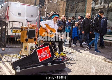 Shoreditch, London, England, UK - April 2019: Junge Gaukler mann straße Musiker Gitarre spielen und singen unter Masse in der Brick Lane, East London Stockfoto