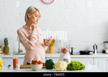 Blonde schwangere Frau mit in der Küche in der Nähe von Cherry Tomaten Smartphone auf Schneidebrett Stockfoto