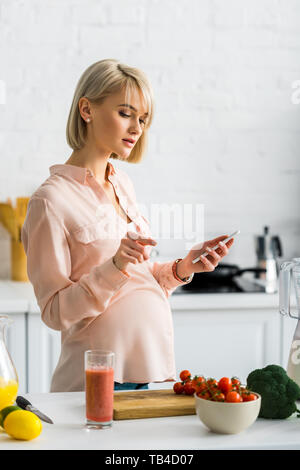 Schwangere Frau mit in der Küche in der Nähe von Cherry Tomaten Smartphone auf Schneidebrett Stockfoto