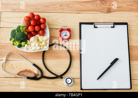 Blick von oben auf die Leere Zwischenablage mit Stift in der Nähe von Stethoskop, Wecker und lecker Gemüse auf Herz-form Platte Stockfoto