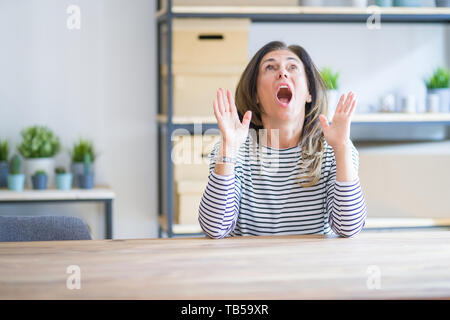 Mittleren Alters ältere Frau am Tisch sitzen zu Hause erstaunt und überrascht mit der Suche und zeigt mit Fingern und erhobenen Armen. Stockfoto