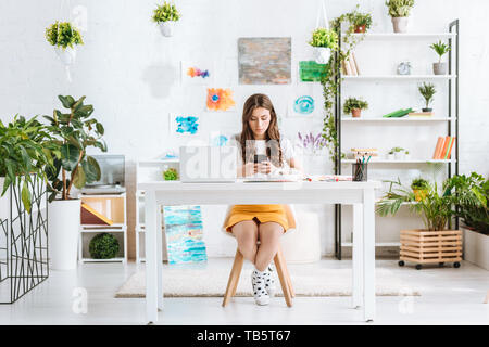 Junge Frau mit Smartphone, während am Schreibtisch mit Laptop im Zimmer zu Hause sitzen Stockfoto