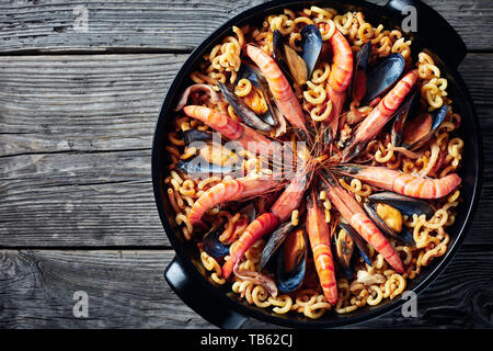 Fideua, einer Paella wie Teller mit Nudeln, Riesengarnelen, weißen Fisch Fleisch, Tintenfische, Muscheln in einem schwarzen Wanne auf einen hölzernen Tisch, Blick von oben, Fla Stockfoto