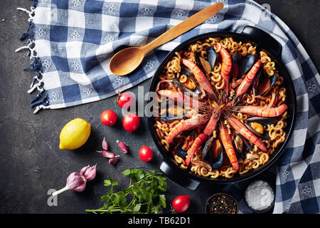 Fideua, einer Paella wie spanisches Gericht mit speziellen Pasta, Riesengarnelen, weißen Fisch Fleisch, Tintenfische, Muscheln in einem schwarzen Wanne auf einen konkreten Tisch mit Zutaten Stockfoto