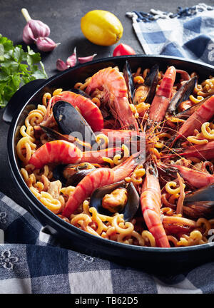 Spanisch Fideua, eine Nudel Paella mit Meeresfrüchten - Riesengarnelen, weißen Fisch Fleisch, Tintenfische, Muscheln in einem schwarzen Wanne auf einen konkreten Tisch mit Zutaten, ve Stockfoto