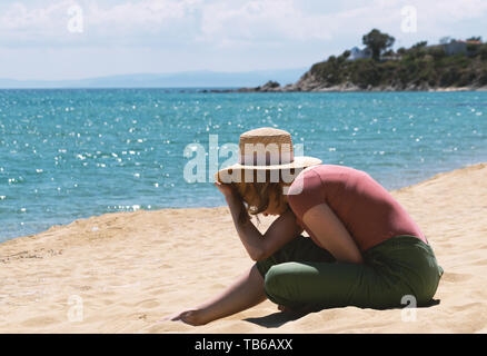 Frau mit casual Outfit sitzt am Sandstrand in der Nähe von blauen Meer und Verstecken von der Heizung Sonne mit Strohhut. Stockfoto