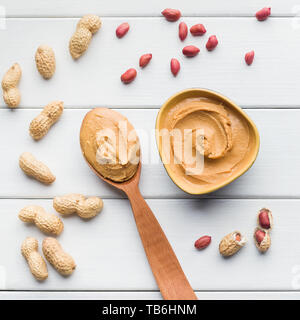 Erdnussbutter in der Schüssel und Löffel und zerstreute Erdnüsse auf weißem Holz- Hintergrund, Ansicht von oben. Artisan Erdnussbutter Konzept Stockfoto