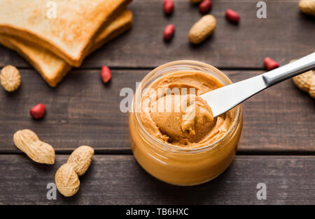 Erdnussbutter im Glas, Toast und zerstreute Erdnüsse auf Holz- Hintergrund. Vegetarische butter Konzept Stockfoto