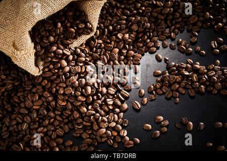 Geröstete Kaffeebohnen mit kleinen Sackleinen Tasche in die Ecke Auslaufen auf einen dunklen Schiefer Oberfläche mit Highlight gesehen hohen Winkel Stockfoto