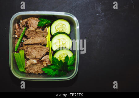 Gekochtes Rindfleisch und frische Gurke in einem kleinen Behälter auf einem schwarzen Hintergrund, Ansicht von oben, aus der Nähe. Gesunde Ernährung, Diät, Fitness. kopieren. Stockfoto