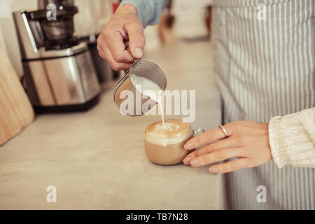 Hinzufügen von Milch. Hand des Barista gießen Milch in eine Tasse Kaffee in der Hand eines Coffee shop Gast. Stockfoto