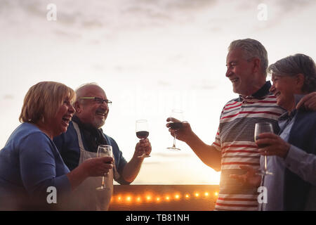 Gerne ältere Freunde trinken Rotwein an Barbecue in Terrasse - reife Menschen jubeln und zusammen lachen bei Sonnenuntergang auf der Dachterrasse Stockfoto