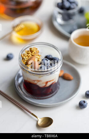 Close up Müsli mit Blaubeeren und Mandel, Marmelade und Joghurt im Glas am grauen Platte auf weißem Hintergrund. Honig, Kaffee, Löffel und Servietten mit b Stockfoto