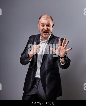 Glücklich, aufgeregt schreien Interpret Mann tanzen und präsentiert die Show holding Mikrofon in der Hand auf grauen Hintergrund. Nahaufnahme Stockfoto