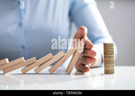 Menschliche Finger stoppen Dominosteine fallen mit gestapelten Münzen auf weißem Schreibtisch Stockfoto