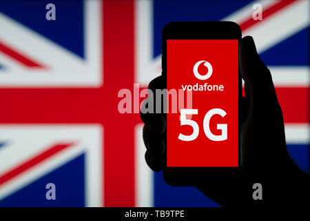 Eine Silhouette einen Mann hält ein Smartphone mit dem Logo von Vodafone und die Buchstaben 5G, vor eine Britische Flagge (nur redaktionelle Nutzung). Stockfoto