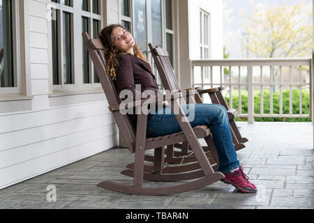 Veranda vor dem Haus mit Jungen glücklich Frau sitzt auf der braunen Schaukelstuhl in der traditionellen amerikanischen Haus Stockfoto