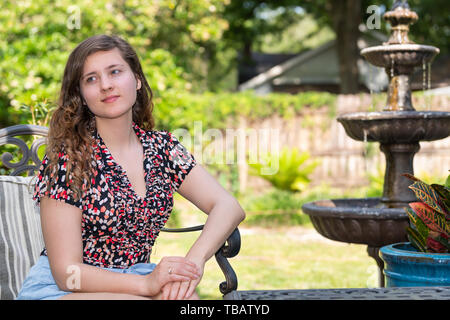 Junge glücklich Frau sitzt auf der Terrasse im freien Stuhl im Frühling Garten im Hinterhof des Hauses lächelnd mit Springbrunnen und Pflanzen Stockfoto