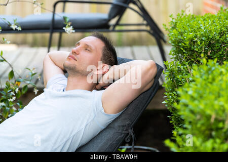 Junge Mann sitzt zurückgelehnt auf Terrasse Lounge Chair schlafen im Freien Frühling Blumen Garten im Hinterhof des Hauses Pflanzen Stockfoto