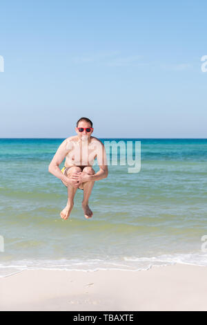 Junge glücklicher Mann springen Bombe in der Luft mit den Armen auf die Fußgelenke mit Ozean Meer, Wellen, die am Horizont im Sommer in Santa Rosa Beach, Florida Stockfoto