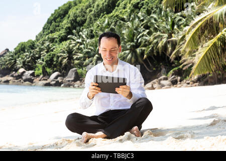Lächelnden jungen Mann sitzt am Sandstrand mit seinen gekreuzten Beinen mit digitalen Tablet Stockfoto