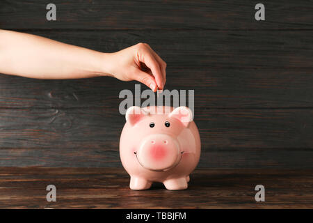 Der Frau die Hand eine Münze in der glücklichen Sparschwein auf Holz Tisch gegen Holz- Hintergrund, Platz für Text. Finanzen, Geld sparen Stockfoto