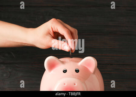 Der Frau die Hand eine Münze in der glücklichen Sparschwein auf Holz Tisch gegen Holz- Hintergrund, Platz für Text. Finanzen, Geld sparen Stockfoto