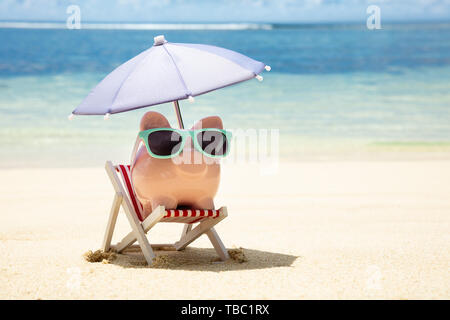 Rosa Sparschwein mit Sonnenbrille und Sonnenschirm auf Liegestuhl auf Sand am Strand Stockfoto