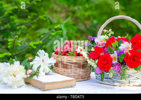 Frühling Sommer noch Leben mit einem Blumenstrauß in einem Korb, Beeren reife Erdbeeren, ein altes Buch und weiße Pfingstrosen auf dem Tisch auf einem natürlichen gre Stockfoto