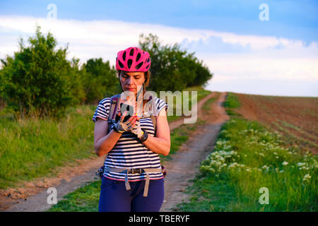 Weibliche Radfahrer Kontrolle ihr Telefon für Richtungen auf eine MTB-Route in der Landschaft, bei Sonnenuntergang, mit einem Feldweg Richtung Horizont als backgrou Stockfoto