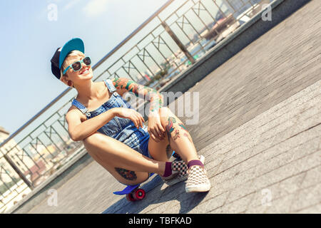 Junge alternative Mädchen skater tragen Cap und Sonnenbrille sitzen auf Penny auf der Straße fröhlich lächelnd beiseite suchen Stockfoto