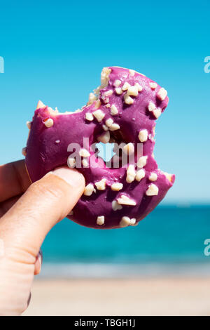 Nahaufnahme eines knabberte appetitlich Donut, beschichtet mit einem violetten Zuckerguss und weiß besprüht, in der Hand des jungen Mannes gegen den blauen Himmel und im OCE Stockfoto