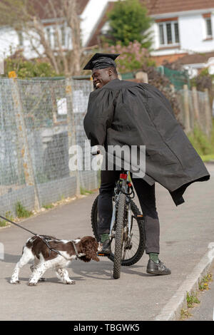 Andover, Hampshire, England, UK. Mai 2019. Eine Universität Student trägt die Kappe und Kleid auf einem Fahrrad. Anschläge auf Bürgersteig zu sagen hallo zu einem Spaniel hund Stockfoto