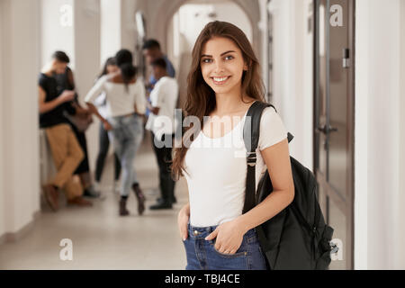 Positivität und glücklich Studentin mit Rucksack auf Korridor von Luxus International University, Mitschüler für den Hintergrund. Brunette Mädchen in Jeans gehen zu Vorlesung, in die Kamera lächeln. Stockfoto