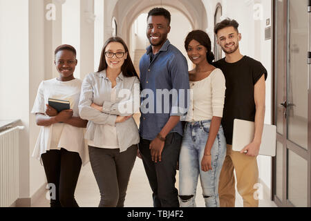 International Gruppe junger Studenten Afrikanische und Kaukasische zusammen stehen und lächelte Flur, sie auch Noten und Rucksack. Hübsche Mädchen und Jungen ruht auf Pause, Zeit miteinander zu verbringen. Stockfoto