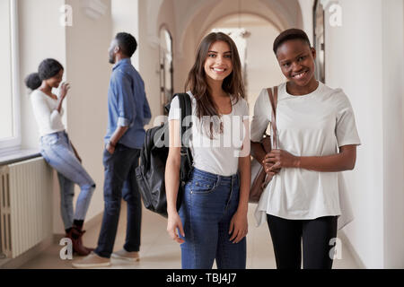 Ziemlich Afrikanische und kaukasische Schüler Mädchen stehen auf Korridor von Universität und lächelnd an der Kamera. Frauen, die Freunde, die in der Internationalen Universität gemeinsam studieren. Stockfoto