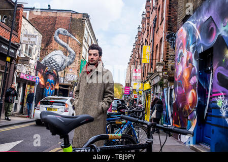 Brick Lane, Shoreditch, London, England, UK - April 2019: Hipster jungen Mann mit langen Winter Mantel in Brick Lane in der Nähe einer Wand in abgedeckt Stockfoto