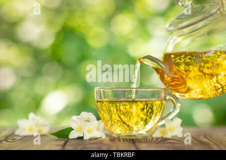 Pflanzliche Jasmin Tee Gießen der Teekanne aus Glas in Schale auf Holztisch mit unscharfen grün Natur Hintergrund Stockfoto