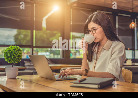 Asiatische arbeitende Frau mit Laptop und trinken Kaffee im Cafe. Die Menschen und Lebensstile Konzept. Technik und Wirtschaft Thema. Freiberufliche und Beruf t Stockfoto