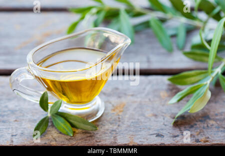 Olivenöl in Glaskanne mit Olivenbaum Zweige auf Holz- Hintergrund. Nahaufnahme Stockfoto