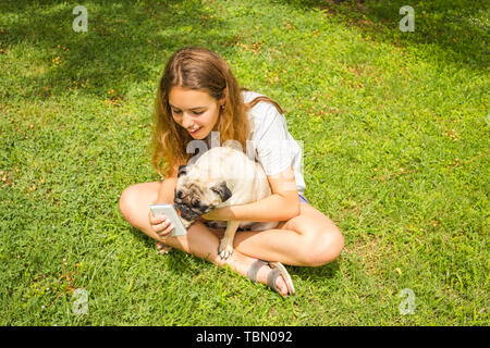 Schöne jugendlich Mädchen beobachten ein lustiges Video auf einem Smartphone mit ihrem Mops Hund im Sommer Park Stockfoto