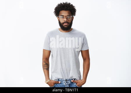 Portrait von ruhig und entspannt unemotional african american reifer Mann mit Bart in Gläsern mit durchbohrten Nasen- und Tattoos halten sich an den Händen in den Taschen suchen Stockfoto