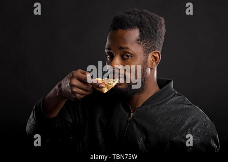 Closeup Portrait der jungen schönen afrikanischen Mann in Schwarz Jacke holding Stück Pizza und zeigen ihn mit Zeigefinger an der Kamera auf der Suche nach dunklen Bac Stockfoto