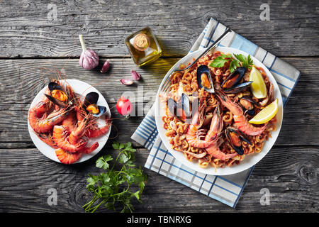 Spanisch Fideua, eine Nudel Paella mit Riesengarnelen, weißen Fisch Fleisch, Tintenfische, Muscheln auf einem weißen Teller auf einem Holztisch mit gebratenen Meeresfrüchten serviert. Stockfoto