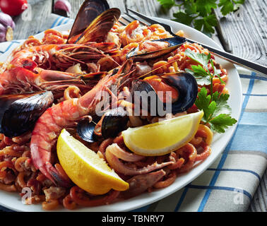 Nahaufnahme der Spanischen Fideua, eine Nudel Paella mit Riesengarnelen, weißen Fisch Fleisch, Tintenfische, Muscheln, serviert auf einem weißen Teller mit einem silbernen Gabel, grau Stockfoto