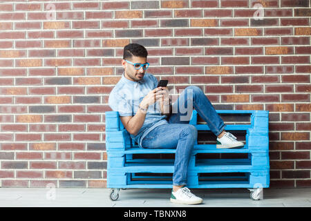In voller Länge Porträt von Ernst hübscher junger bärtiger Mann im lässigen Stil und Brillen sitzen auf Blau Holzpalette, Holding sein Smartphone und Re Stockfoto