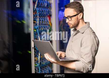 Ernste nachdenkliche hübscher junger server Engineer in Gläsern stehen in der Datenbank und mit Laptop während der Fehlerbehebung Problem mit Netzwerk Stockfoto