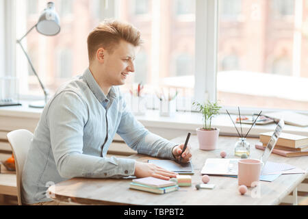 Lächelnd selbstbewussten jungen männlichen Designer im casual Shirt sitzt am Schreibtisch und mit Grafiktablett während der Ausarbeitung Creative Design für mobile App Stockfoto